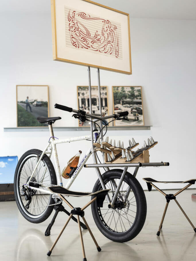 Ein Fahrrad ist zu sehen, darauf befestigt ist eine Metallkonstruktion die eine gerahmte Kalligrafie auf einer Partitur trägt. Dafür ist eine Skulptur zu sehen, die aus Spritzköpfen besteht. Daneben steht ein dreibeiniger Hocker.