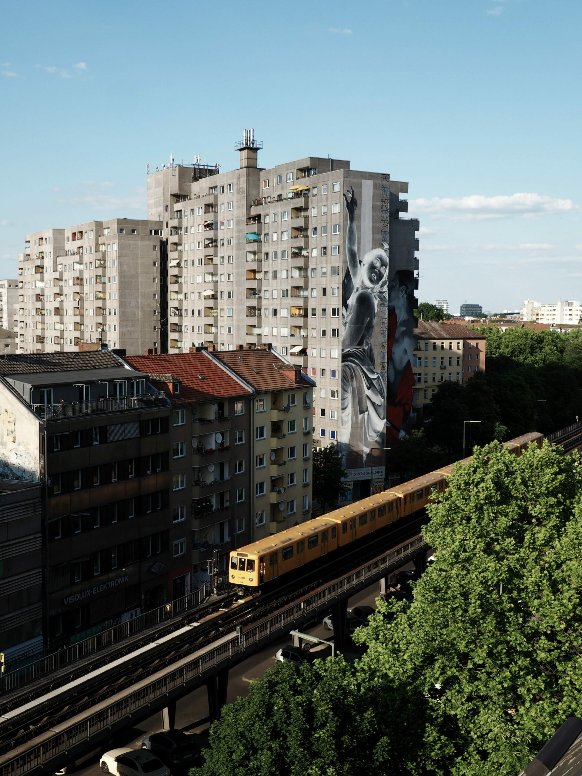 Ein Bild der Berliner U-Bahn auf einer überirdisch verlaufenden Linie (U2), dahinter ein Plattenbau mit einem Mural, dass eine invertierte Frau zeigt. 