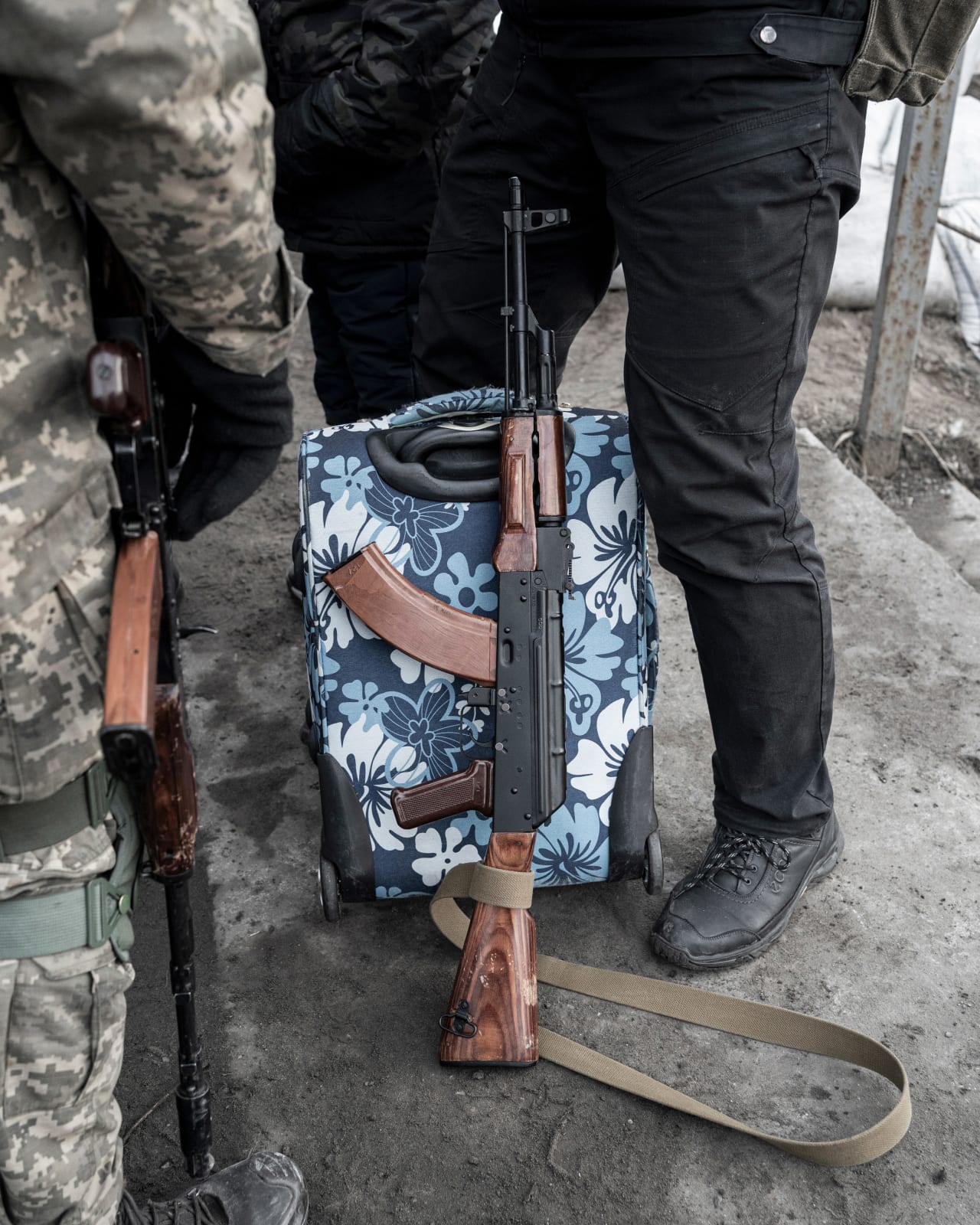 A gun leaning against a bright suitcase, left and right the legs of soldiers. 