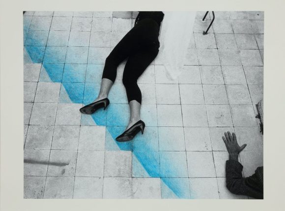 A woman in heels laying on tiled floor, blue color is painted in a stairway style and a male hand is touching the floor from the right bottom corner.