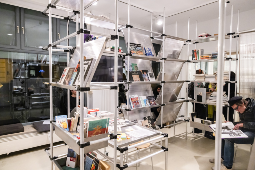 Die Bücherei des  C& Center of Unfinished Business der ifa-Galerie Berlin. Zu sehen sind Menschen, die sich mit den Büchern befassen und zwischen den Regalen lesen oder sich unterhalten.