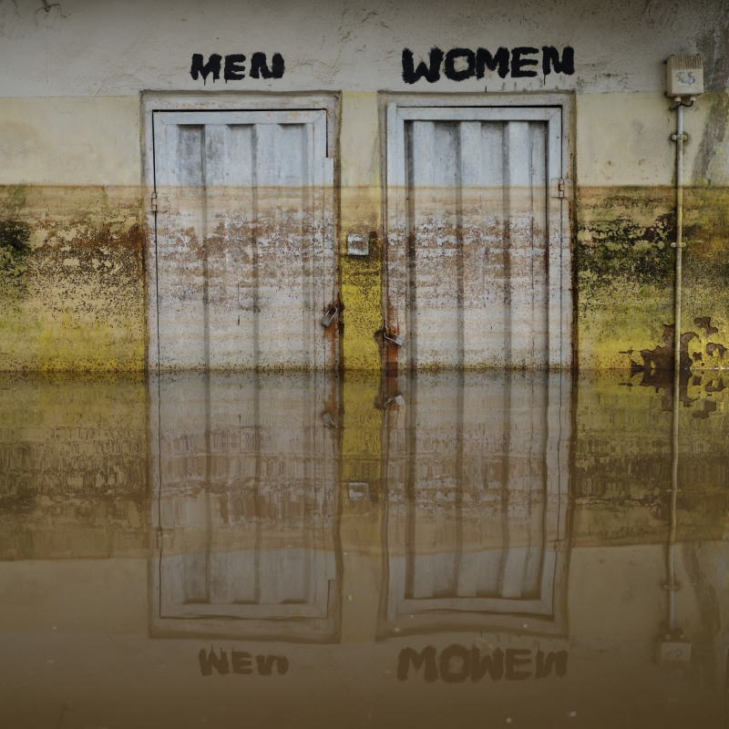 Eine alte Fabrikhalle mit zwei Türen über denen jeweils "Men" und "Woman" steht. Im Vordergrund spiegelt sich die Szenerie im Wasser wieder.