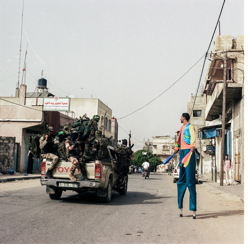 Auf der linken Seite ist ein übervoller Pick-Up Truck mit Soldaten zu sehen, auf der rechten Seite ein Jongleur auf Stelzen in bunten Gewändern. Die Szene wurde in Afghanistan aufgenommen.
