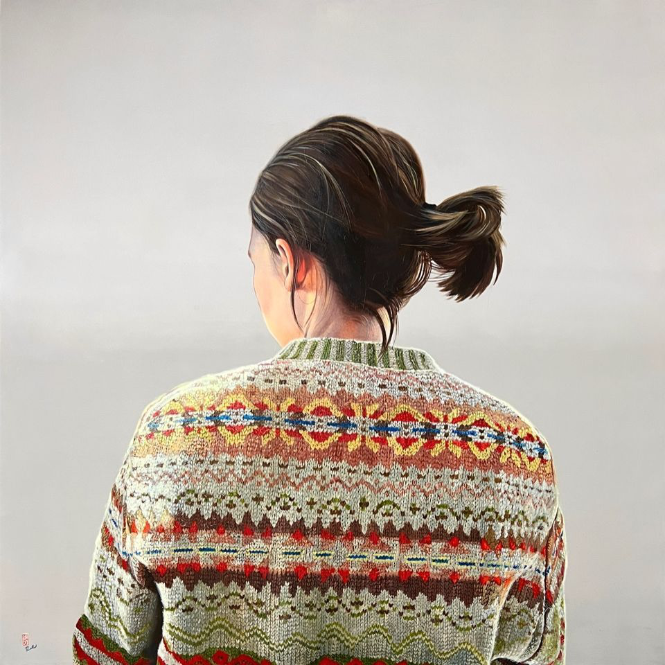 Zu sehen ist das Portrait einer jungen Frau in der Rückansicht vor einem hellgrauen Hintergrund. Sie trägt einen gestrickten Wollpullover mit buntem Zopfmuster und ihr Haar ist hochgebunden. 