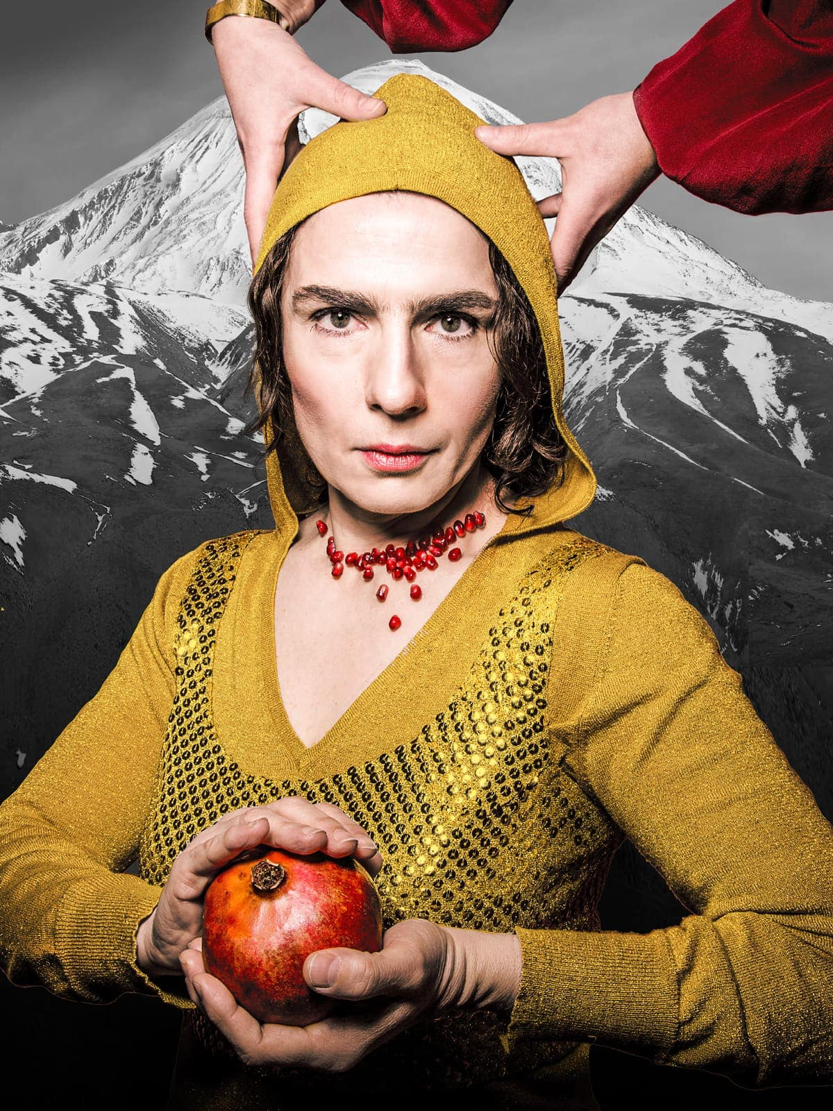 Portrait von Aurora Mardiganian in einem gelben Hoodie und geschmückt von einer Granatapfelsamenkette. Zwei Hände fassen der dunkelhaarigen Frau, die einen Granatapfel in der Hand trägt an den Kopf. Ihr Blick ist entschlossen und eindringlich, die Lippen blass rosa und die Züge ihres Gesicht angespannt. Dahinter ist eine Bergkette zu sehen.