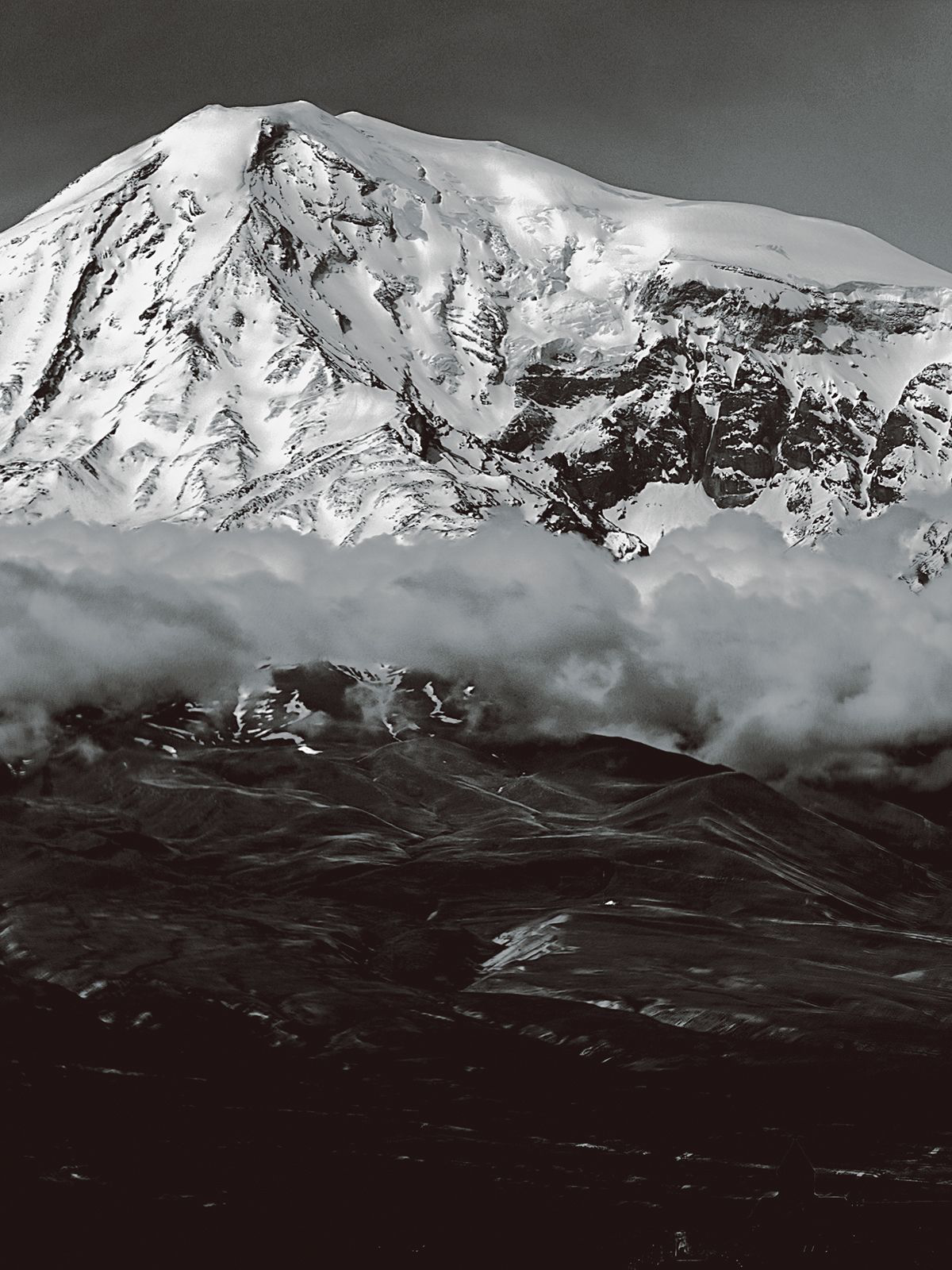 Zu sehen ist eine armenische Berglandschaft. Die Mitte des Bildes ist durch eine Wolkendecke geteilt. Dahinter ragt ein schneebedeckter Berg auf. Im Vordergrund ist eine schwarze Landschaft zu sehen.