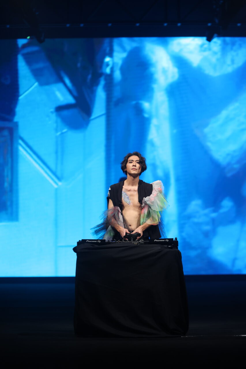 Eine Person mit kurzen Haaren steht hinter einem DJ-Pult auf einer schwach beleuchteten Bühne und trägt das #DAMUR SS25_00011 – ein durchsichtiges, farbenfrohes Outfit. Im Hintergrund zeigt ein großer blauer Bildschirm abstrakte Bilder.