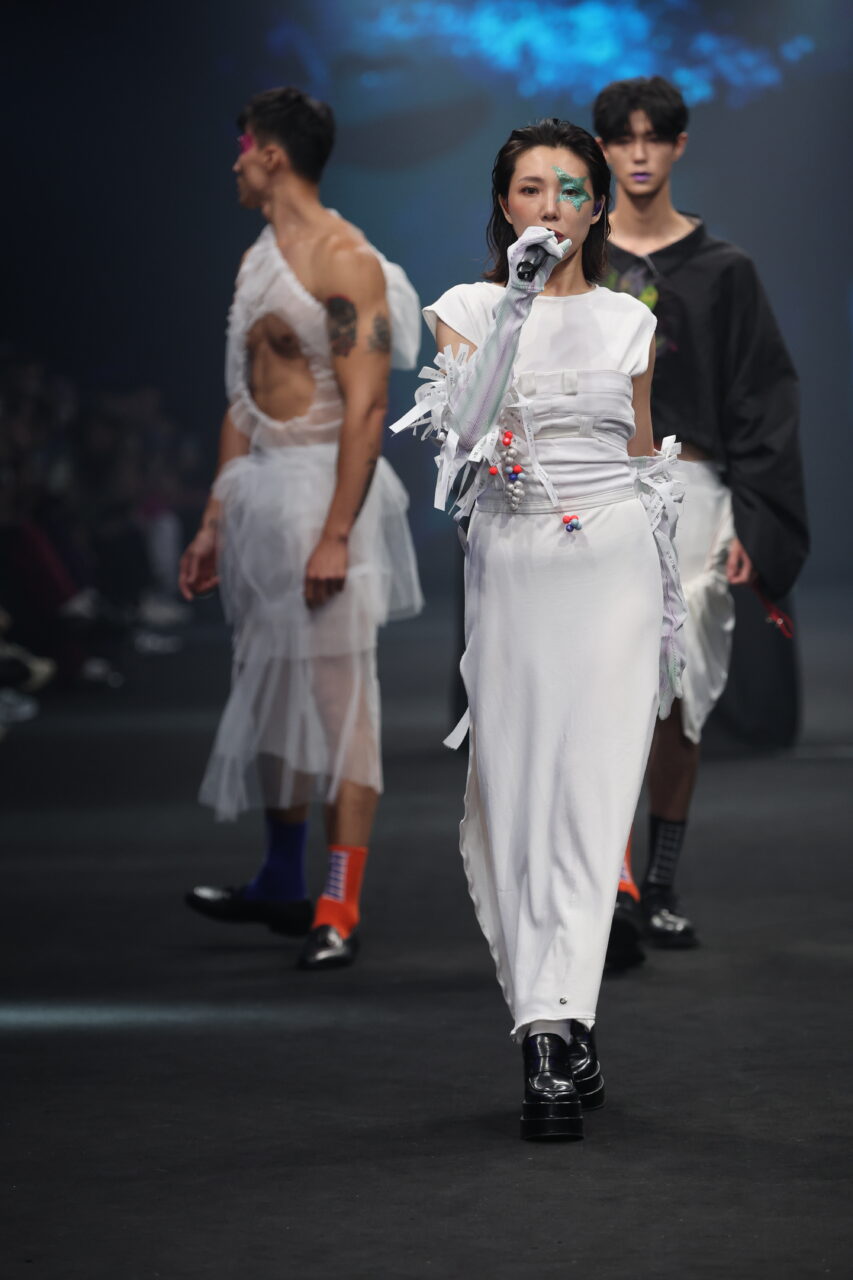 Models laufen in avantgardistischer Mode über einen schwach beleuchteten Laufsteg und präsentieren die Kollektion #DAMUR SS25_00041 mit weißen und schwarzen Outfits mit einzigartigen Designs und Accessoires.