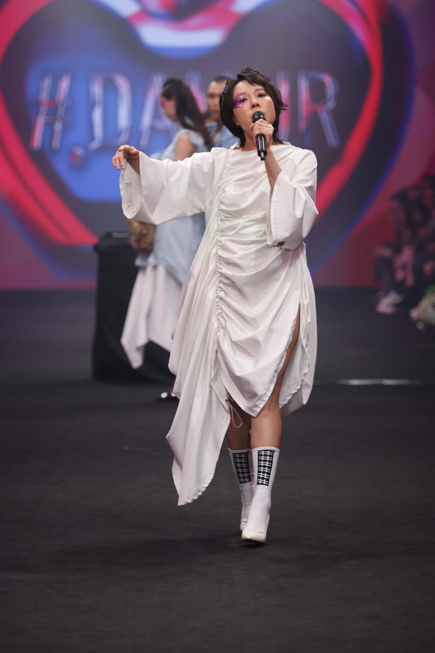 Vor einer „#DAZZLE“-Kulisse und mit Publikum im Hintergrund läuft ein Model den Laufsteg entlang und präsentiert das Ensemble #DAMUR SS25_00185, komplett mit weißem Kleid, Stiefeln und einem Mikrofon in der Hand.