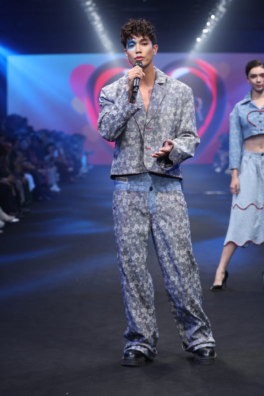 Ein Model läuft über den Laufsteg und trägt das #DAMUR SS25_00292, ein Jeans-Outfit mit Blumenmuster, und hält ein Mikrofon in der Hand. Im Hintergrund ist ein weiteres Model zu sehen.