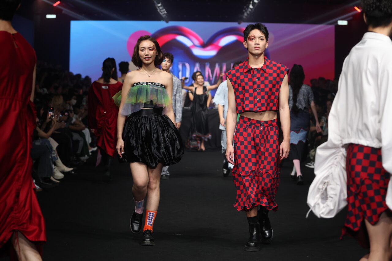 Models laufen in der Kleidung #DAMUR SS25_00484, in farbenfrohen und vielseitigen Outfits, über einen Laufsteg, während im Hintergrund eine Menschenmenge zusieht.