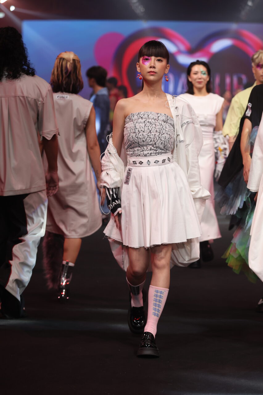 Models laufen in lebhaften und stylischen Outfits über den Laufsteg. Im Mittelpunkt steht ein Model, das #DAMUR SS25_00505 trägt, mit einem weißen Lagenrock und Top.