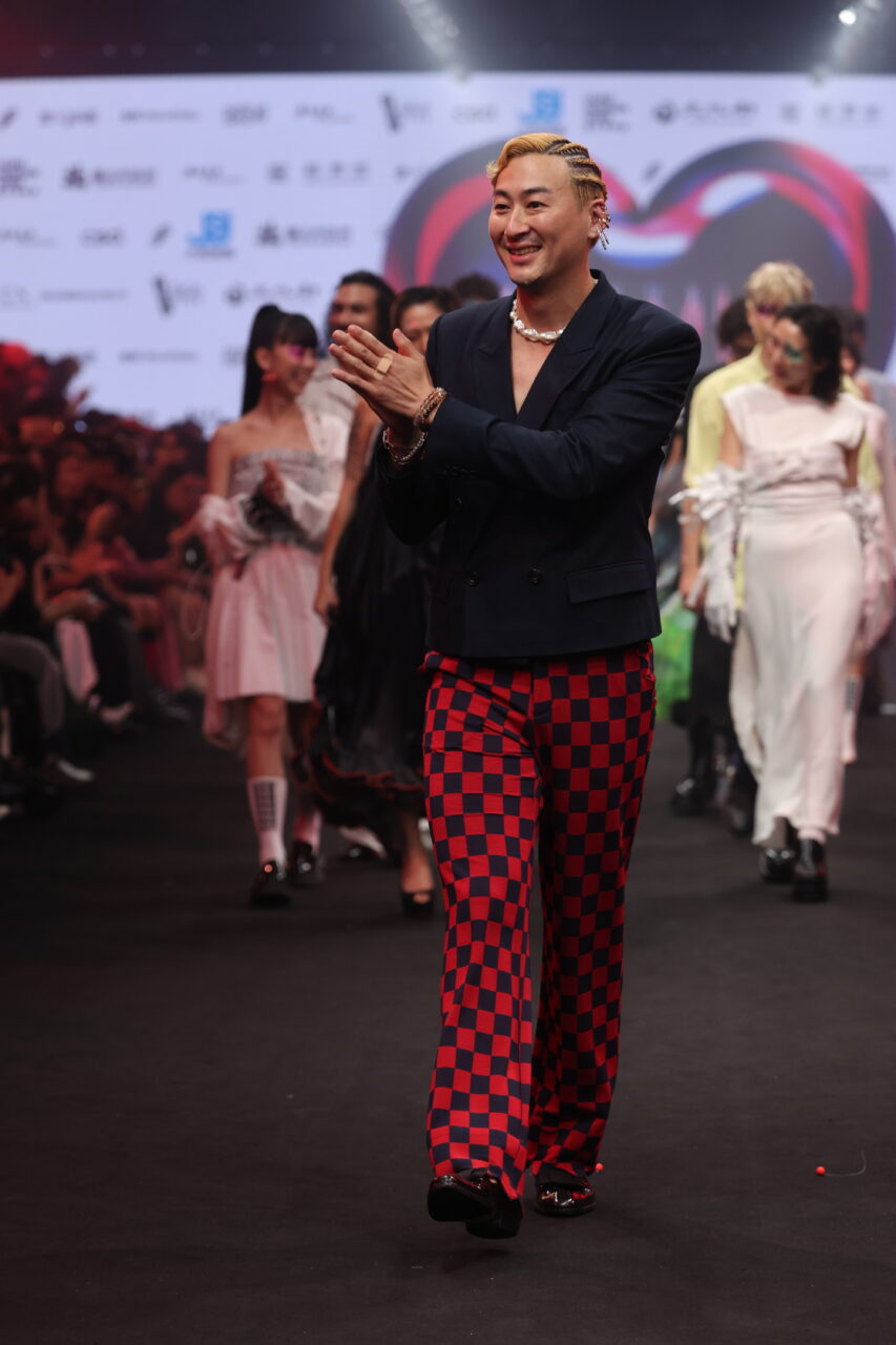 Eine Person, die die karierte Hose #DAMUR SS25_00568 trägt, schreitet über einen Laufsteg und führt eine Gruppe von Models an.