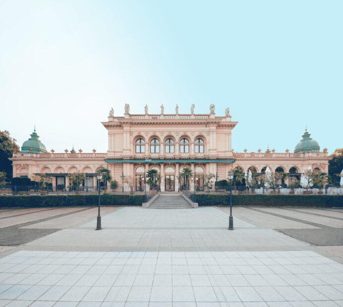 Der historische Kursalon Wien, mit freundlicher Genehmigung des Kursalon Wien, präsentiert kunstvolle Architektur mit Säulen, Statuen und einer Reihe von Bogenfenstern. Das Gebäude ist wunderschön vor einem klaren blauen Himmel mit einem gepflasterten Bereich im Vordergrund zu sehen.